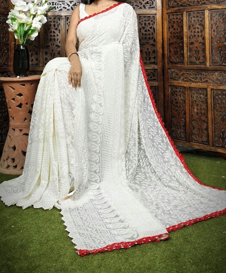 May include: A white saree with intricate embroidery and a red border. The saree is draped over a person, showcasing the delicate floral patterns and detailed design. The saree is made of a lightweight fabric.
