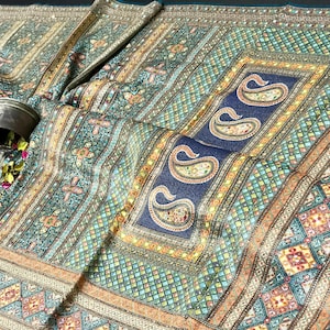 May include: An ornate textile featuring a detailed pattern in teal, gold, and navy blue. The design incorporates paisley motifs and geometric shapes. A small metal container is visible, adding a contrasting element to the fabric's intricate design.