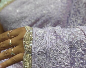以下が含まれることがあります： 淡いラベンダー色のサリーで、複雑な白い花柄の刺繍と繊細なスパンコールの装飾が施されています。生地の縁はスカラップ状になっており、ゴールドトーンのメタリック糸と小さな反射スパンコールが布地の端に沿ってあしらわれています。
