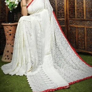 May include: A white saree with intricate floral embroidery and a red border. The saree is draped, showcasing the detailed patterns and textures. The red border adds a contrasting accent to the overall design. The saree is made of a lightweight fabric.