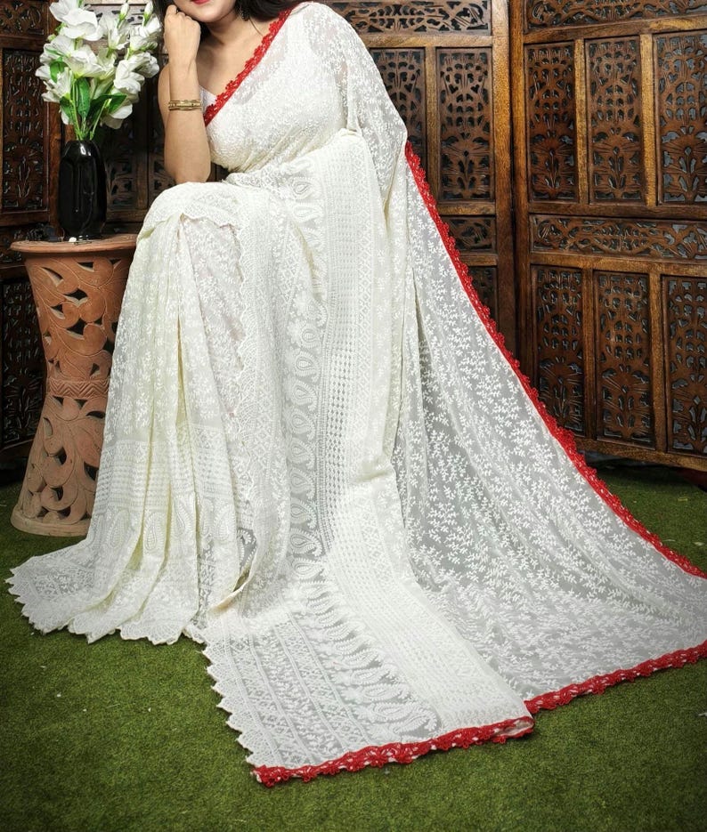 May include: A white saree with intricate floral embroidery and a red border. The saree is draped, showcasing the detailed patterns and textures. The red border adds a contrasting accent to the overall design. The saree is made of a lightweight fabric.
