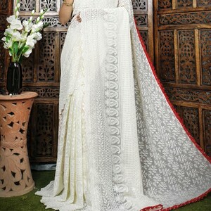 May include: A white saree with intricate embroidery and a red border. The saree features detailed patterns and a flowing design, showcasing traditional craftsmanship. The fabric appears lightweight and delicate, suitable for special occasions.