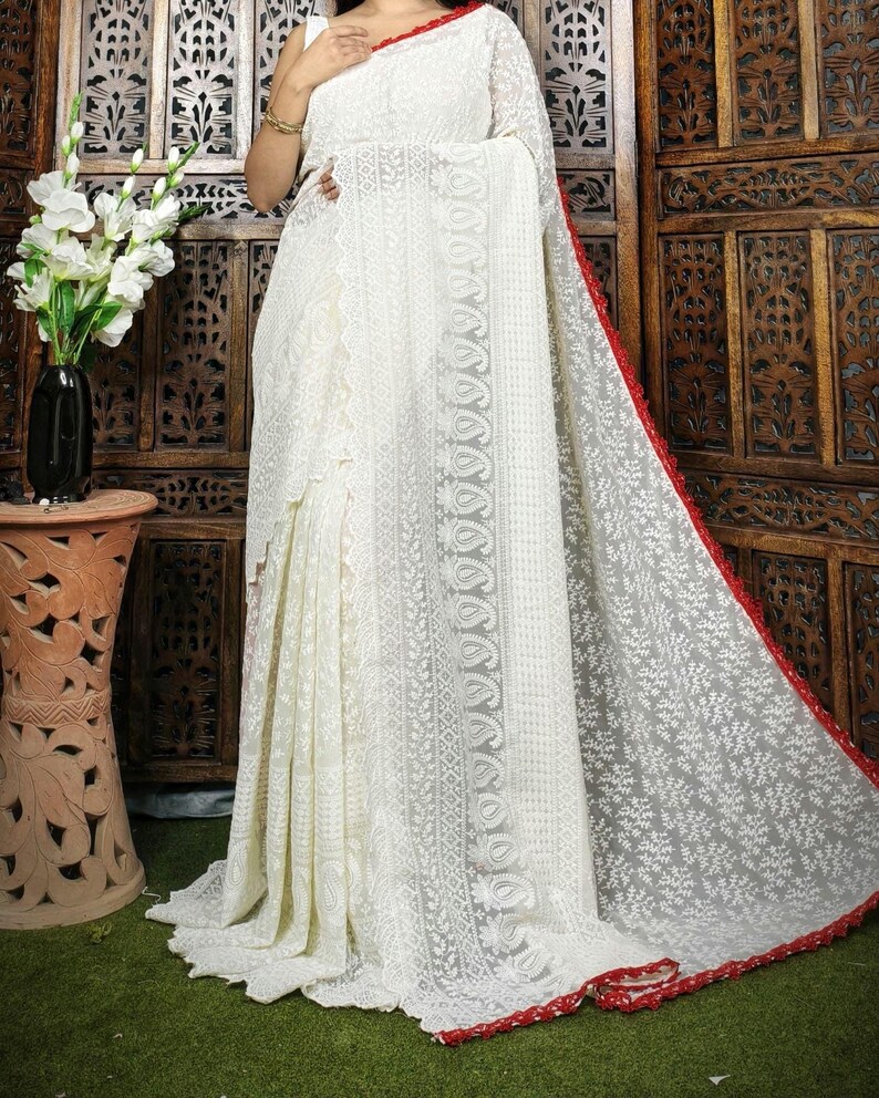May include: A white saree with intricate embroidery and a red border. The saree features detailed patterns and a flowing design, showcasing traditional craftsmanship. The fabric appears lightweight and delicate, suitable for special occasions.
