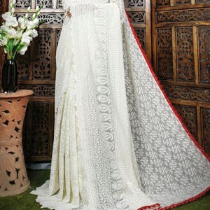 May include: A white saree with intricate floral embroidery. The saree features a red border and a delicate, sheer fabric. The design includes detailed patterns and a flowing drape, showcasing traditional craftsmanship.