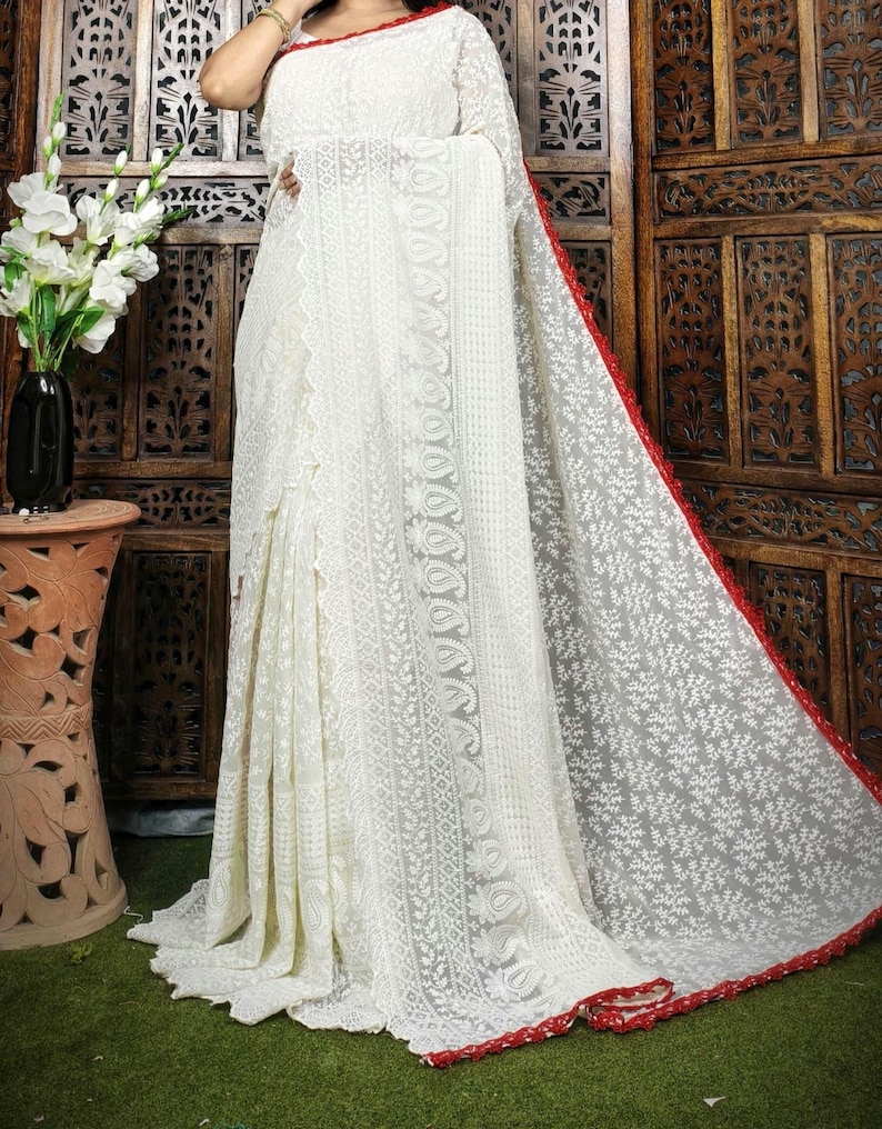 May include: A white saree with intricate floral embroidery. The saree features a red border and a delicate, sheer fabric. The design includes detailed patterns and a flowing drape, showcasing traditional craftsmanship.