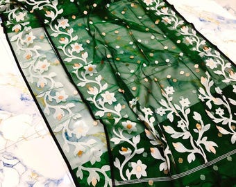 Puede incluir: Una tela verde esmeralda transparente con bordado floral blanco y un borde negro. El diseño presenta delicadas flores y hojas blancas, con pequeños acentos dorados dispersos por la superficie. La tela está drapeada, mostrando su textura fluida.