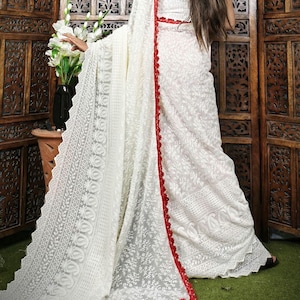 May include: A white saree with intricate floral embroidery and a red border. The saree is draped over a person, showcasing the detailed patterns and textures of the fabric. The background features wooden panels and a vase of white flowers.