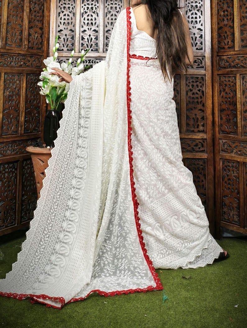 May include: A white saree with intricate floral embroidery and a red border. The saree is draped over a person, showcasing the detailed patterns and textures of the fabric. The background features wooden panels and a vase of white flowers.