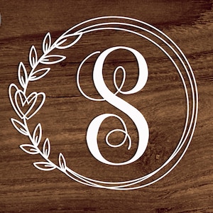 May include: White outline of a wreath with leaves and hearts surrounding the letter S in a double circle frame on a brown wood background.