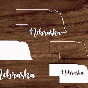 May include: Four white outlines of the state of Nebraska with the word "Nebraska" written in cursive underneath each outline. The outlines are on a brown wood background. The file types available are SVG, PNG, DXF, and PDF.