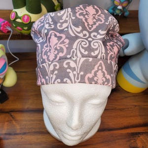 May include: A gray and pink patterned hat is displayed on a white mannequin head. The hat features a repeating floral design in pink and white against a gray background. The hat has a folded brim.