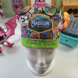 May include: A green and multi-colored fabric scrub hat with a variety of national park patches. The hat features a large patch that says "Oregon Crater Lake".