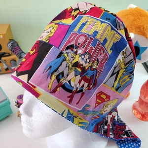 May include: A colorful fabric head wrap with a comic book pattern featuring female superheroes. The wrap has a blue background with pink, yellow, and red accents. The text "Femme Power" is printed in white on the wrap.