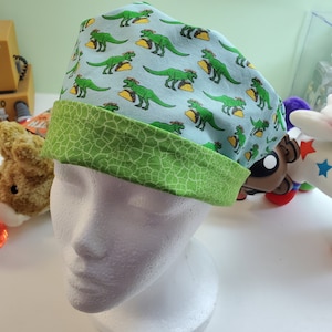 May include: A light blue surgical scrub cap featuring a pattern of green dinosaurs holding tacos. The cap has a green, textured band. This fun, novelty cap is designed for medical professionals.