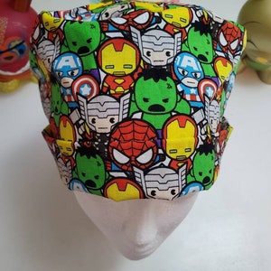 May include: A colorful fabric scrub hat with a pattern of cartoon superheroes, including Iron Man, Captain America, Thor, Hulk, and Spiderman.