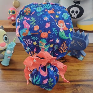 May include: A blue fabric scrunchie with a mermaid and sea life pattern. The scrunchie is tied with a coral-colored ribbon.