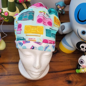 May include: A light blue scrub cap featuring a repeating pattern of retro boomboxes in pink, yellow, and teal. The cap is displayed on a white mannequin head. The words "HOT BEATS" are visible on the cap.
