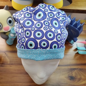 May include: A blue and white surgical scrub hat with a pattern of blue evil eyes on a white background. The hat has a blue and white swirl pattern trim.