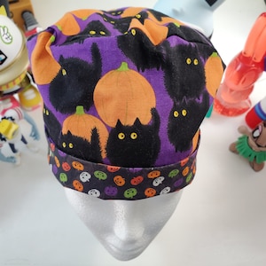 May include: A Halloween-themed scrub cap featuring a purple background with black cats and orange pumpkins. The brim has a pattern of pumpkins and skulls. This cap is designed for medical professionals or anyone wanting a festive touch.