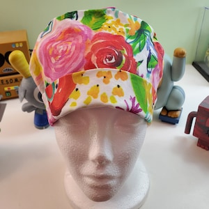 Puede incluir: Un gorro quirúrgico con estampado floral. Presenta grandes rosas rosas y rojas, acentos amarillos y naranjas, y hojas verdes sobre un fondo blanco. El borde es blanco con detalles florales amarillos. Ideal para profesionales de la salud.