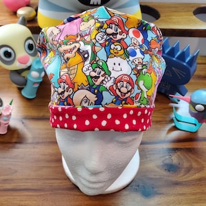 May include: A colorful chef's hat featuring a vibrant print of various characters from the Super Mario video game series. The hat has a red band with white polka dots. The hat is displayed on a white mannequin head.