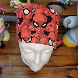 May include: A red and black Spider-Man patterned scrub cap. The cap is displayed on a white mannequin head. The fabric features a repeating pattern of Spider-Man's face. The background includes various figurines and a wooden surface.