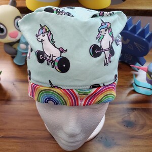 May include: A turquoise blue scrub hat with a rainbow trim. The hat features a repeating pattern of white unicorns lifting barbells.