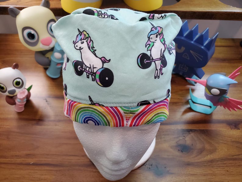May include: A turquoise blue scrub hat with a rainbow trim. The hat features a repeating pattern of white unicorns lifting barbells.