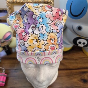 May include: A colorful fabric hat featuring a pattern of Care Bears in various colors. The hat has a white brim with rainbow and heart designs. The hat is placed on a white mannequin head.