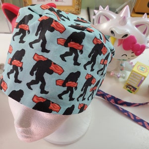 May include: A blue and white scrub hat with a pattern of Bigfoot carrying bacon. The hat has a tie on the back.