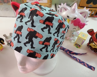 May include: A blue and white scrub hat with a pattern of Bigfoot carrying bacon. The hat has a tie on the back.