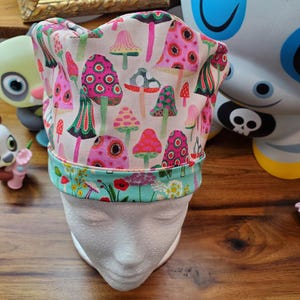 May include: A white scrub hat with a colorful mushroom print. The hat has a turquoise trim and a pink and white floral pattern on the trim.