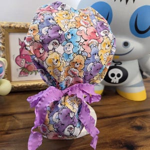 May include: A colorful scrub cap featuring a pattern of Care Bears in various colors. The cap is tied with a lavender ribbon. The background includes a framed picture and a decorative figurine.
