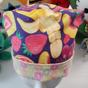 May include: A purple fabric hat with a colorful fruit print. The hat has a white band with a citrus print. The fruits include bananas, strawberries, peaches, raspberries, and grapes.