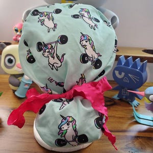 May include: A turquoise blue scrub hat with a pink ribbon tie. The hat has a repeating pattern of white unicorns lifting barbells. The text "Be" and "Un" are visible on the hat.