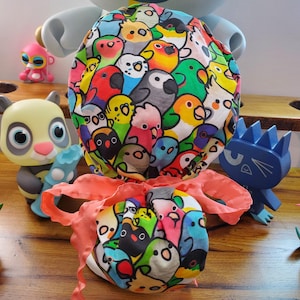 Puede incluir: Un gorro de quirófano de tela colorido con un patrón de pájaros de dibujos animados. El gorro está atado con una cinta rosa y está rodeado de varios juguetes coloridos.