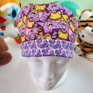 May include: A purple and yellow patterned scrub cap featuring a cartoon character design. The cap has a geometric patterned band in shades of purple, pink, and white. The cap is displayed on a white mannequin head.