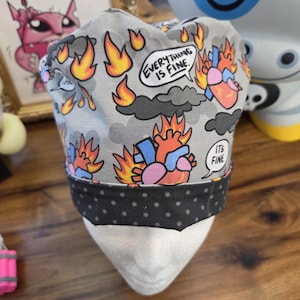 May include: A gray scrub cap with a black polka dot band. The cap features a repeating pattern of cartoon hearts on fire, flames, and speech bubbles with the phrases "Everything is Fine" and "It's Fine."