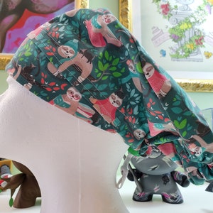 May include: A green, pink, and brown scrub hat with a pattern of sloths and leaves. The hat has a tie at the back.