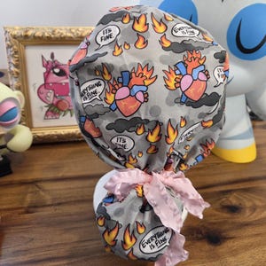 May include: A gray surgical scrub cap with a repeating pattern of flaming hearts, flames, and speech bubbles that say "It's Fine" and "Everything is Fine." The cap is tied with a pink bow.