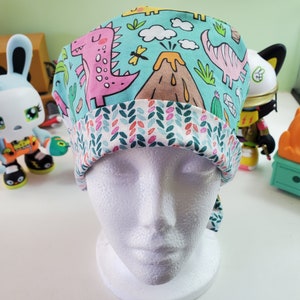 May include: A turquoise blue scrub hat with a dinosaur and volcano print. The hat has a white and green floral print on the inside.