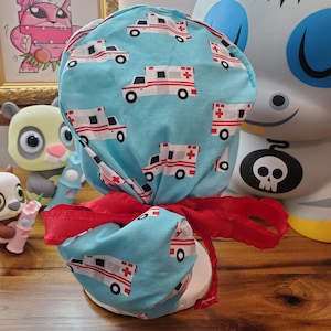 May include: A blue and white patterned scrub hat with a red ribbon tied around it. The pattern features a repeating design of white ambulances on a light blue background.