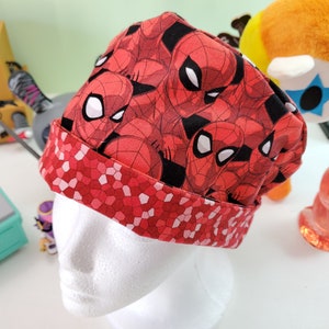 May include: Red and black fabric hat with a repeating pattern of Spiderman's face. The hat has a red and white geometric pattern on the bottom edge.