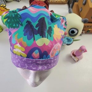 May include: A colorful surgical scrub hat with a purple trim. The hat features a whimsical pattern of trees, hills, and clouds.
