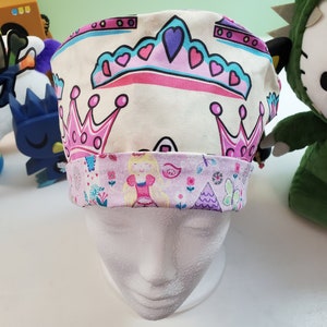 May include: A white scrub hat with a pink and blue princess crown design. The hat has a pink and white floral print on the bottom edge.