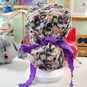 May include: A purple and white scrub hat with a colorful print of cartoon characters. The hat has a purple ribbon tied around it.