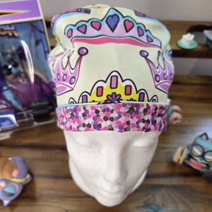 May include: A scrub cap featuring a light yellow background with a repeating pattern of pink, purple, and yellow crowns. The bottom band of the cap has a contrasting print of small, dense purple, pink, and grey dots on a dark background.