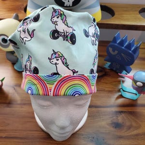 May include: A turquoise blue scrub hat with a rainbow trim. The hat features a repeating pattern of white unicorns lifting weights.