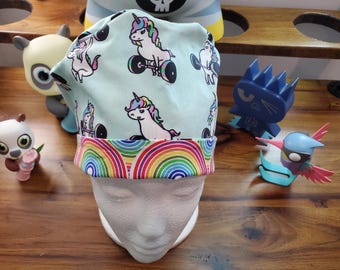 May include: A turquoise blue scrub hat with a rainbow trim. The hat features a repeating pattern of white unicorns lifting weights.