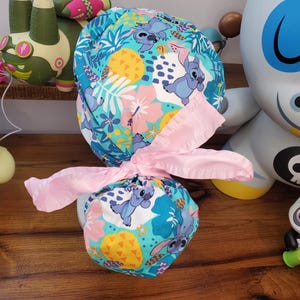May include: A blue and white scrub cap featuring a colorful pattern of the character Stitch, pineapples, and tropical flowers. The cap is tied with a pink bow. The cap is likely for medical professionals or those in the healthcare industry.
